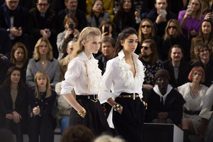 Two models walking in white blouses with ruffles and black shorts at the Chanel fall 2020 show