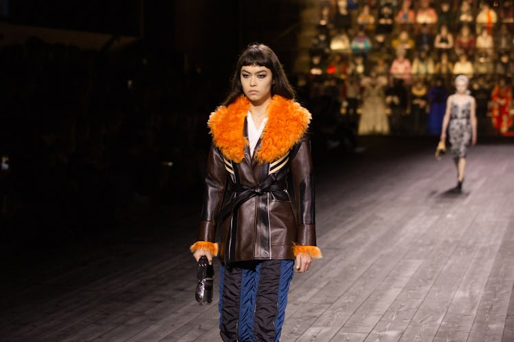 A model in a brown jacket with orange fur collar and blue-black skirt at the Louis Vuitton fall 2020...