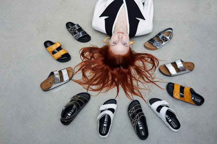 Ginger haired model laying on the ground while Birkenstock sandals and slippers are around her head