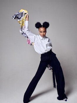 Genneya Walton posing while wearing a white shirt and black pants and holding a colorful scarf