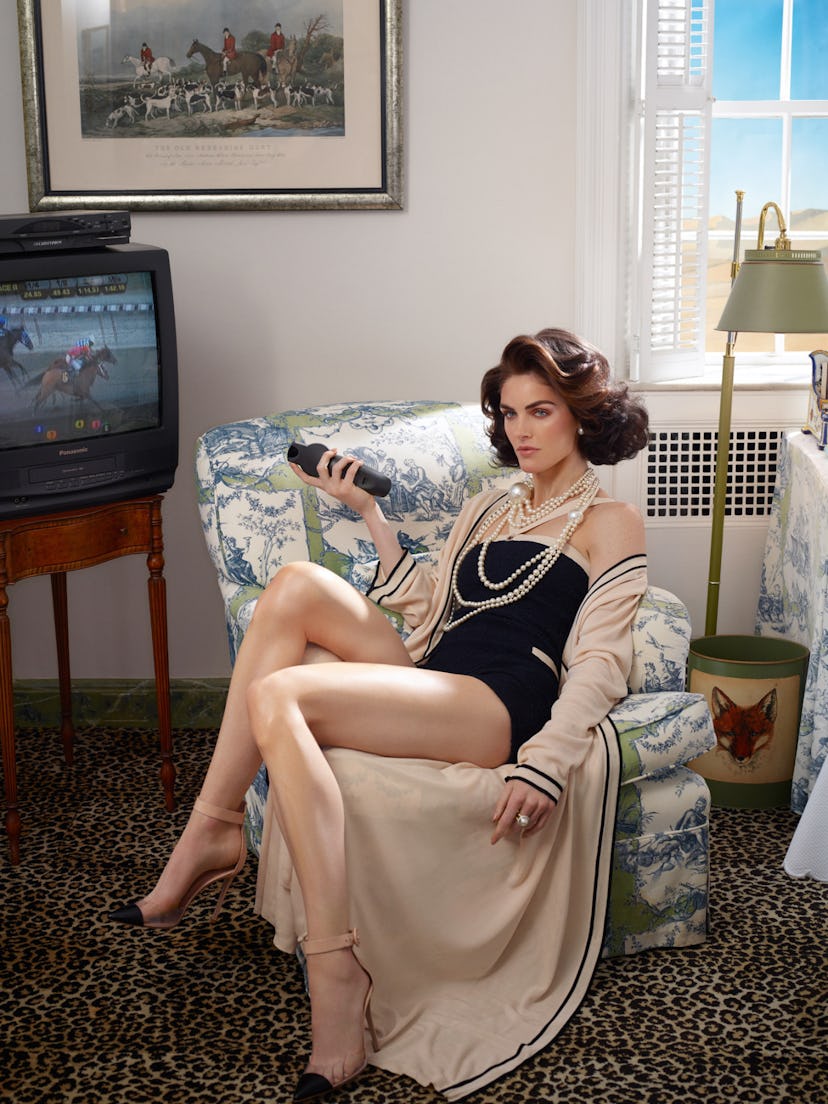 A model in a black bodysuit, and a beige cape with several pearl necklaces sitting in an armchair, h...