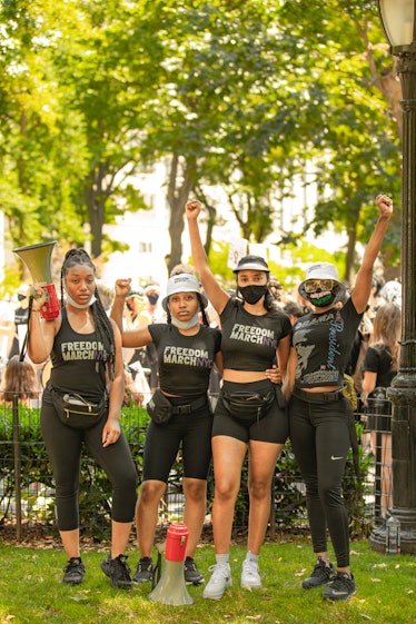 Four women standing with their arms up in black tops with the text 'Freedom March NYC' on the 4th of...