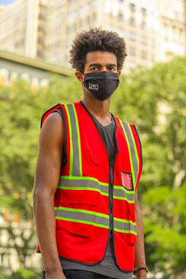 A man standing in a grey top, a red-green reflective vest and a black face mask on the 4th of July