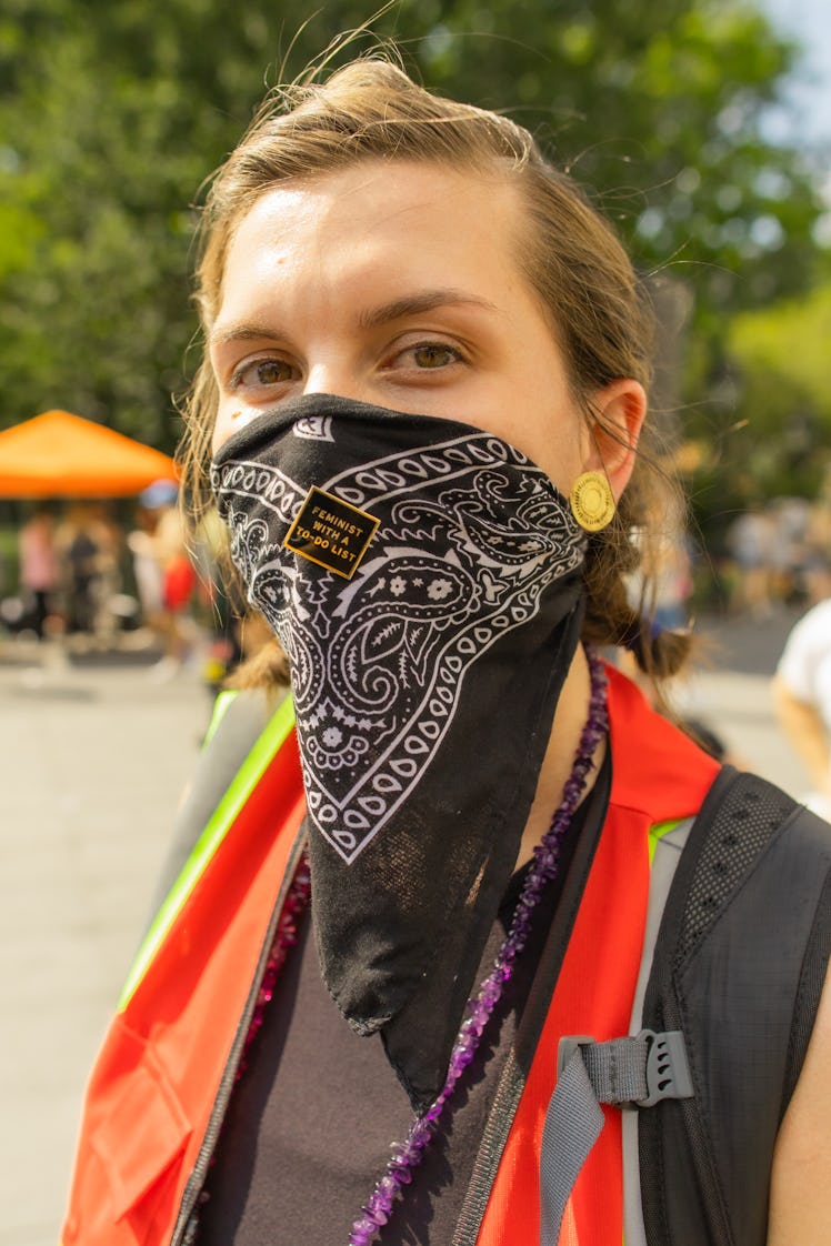 A woman in a black top, red-green reflective jacket and a black bandana covering her face on the 4th...