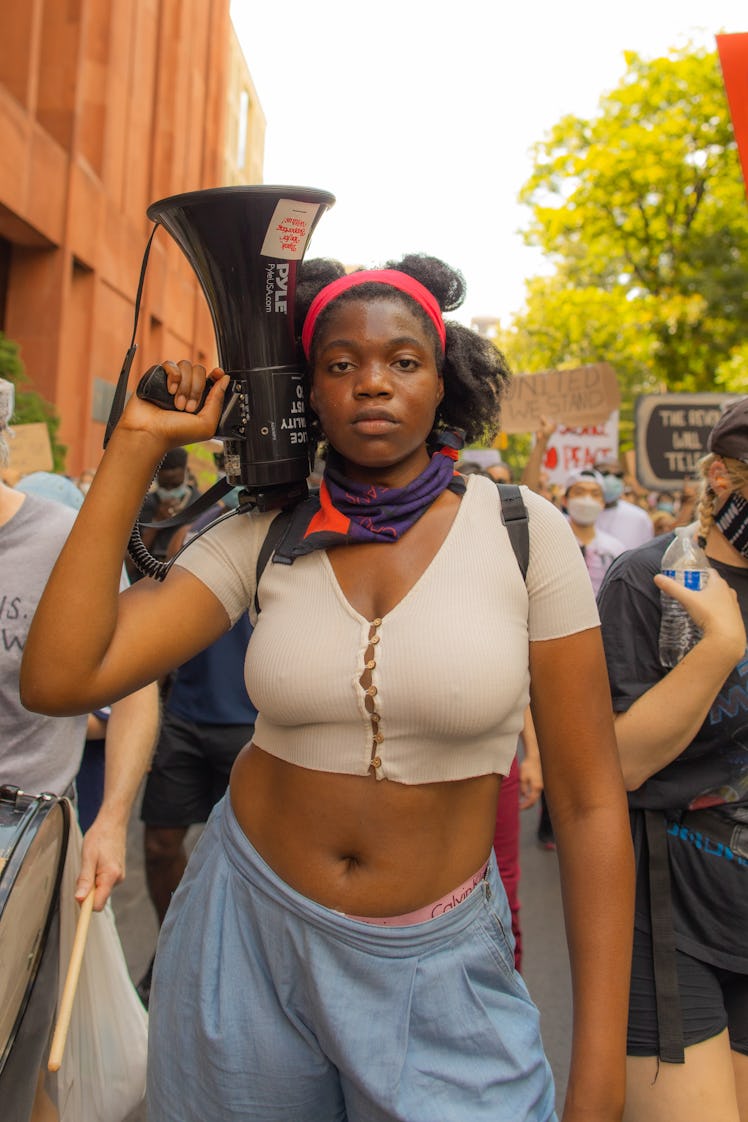 A woman in a beige crop top, blue trousers and purple bandana holding a megaphone on the 4th of July