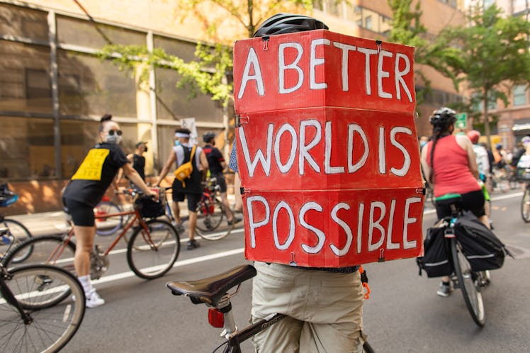 A bicyclist holding a red protest sign with the text 'A BETTER WORLD IS POSSIBLE' on the 4th of July