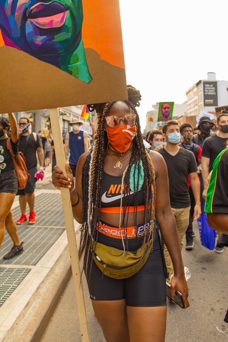 A woman in a black Nike top and shorts and orange face mask holding a painting with George Floyd on ...