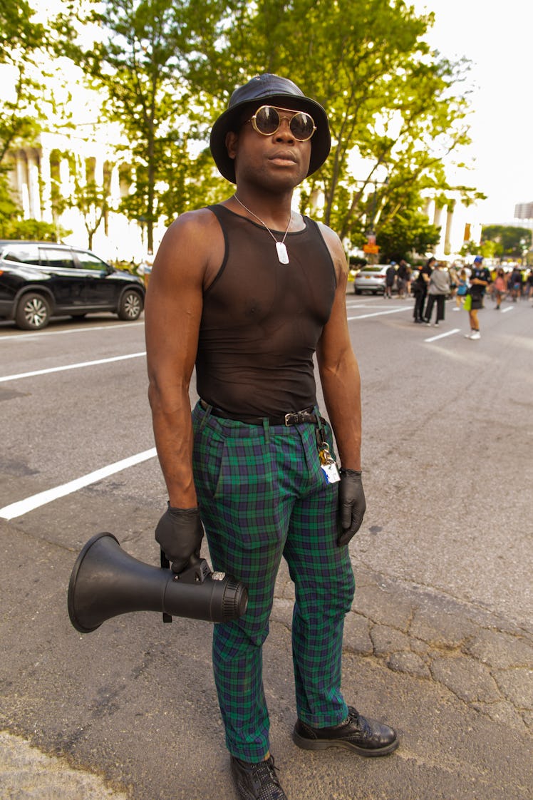 A man in a black tulle top and green checked trousers and a black bucket hat holding a megaphone on ...