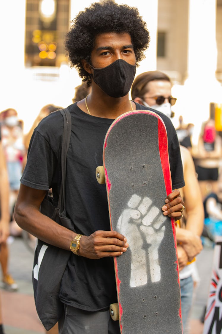 A man in a black shirt, trousers and face mask holding a skateboard with a BLM sign on it on the 4th...