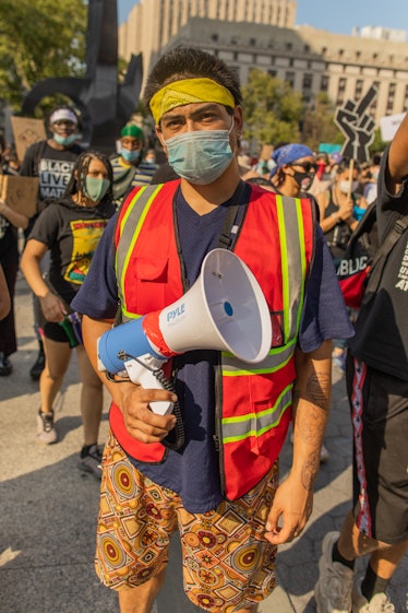 A man in a blue shirt, red-green vest, orange floral shorts, grey face mask and yellow headband hold...