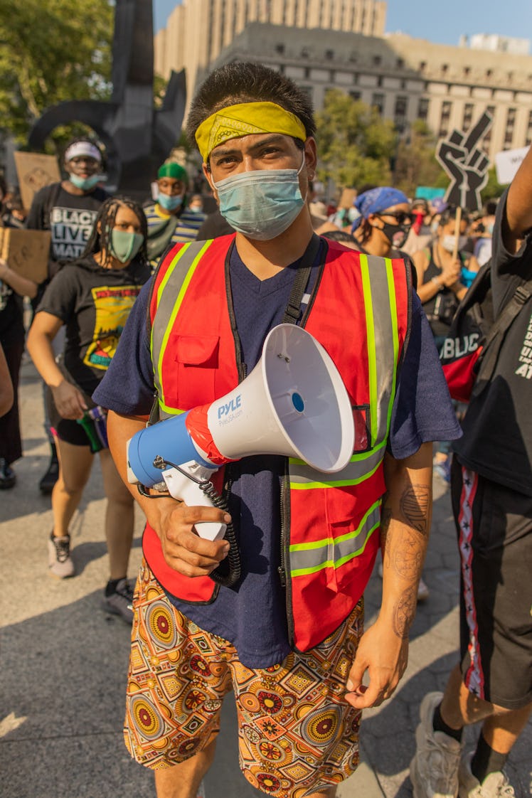 A man in a blue shirt, red-green vest, orange floral shorts, grey face mask and yellow headband hold...