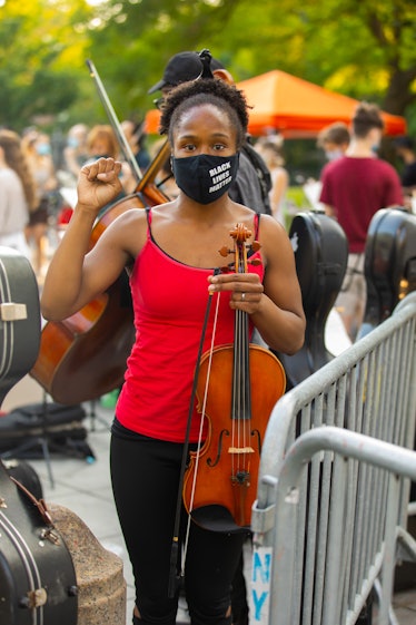 A woman in a red shirt, black pants and a face mask holding a violin on the 4th of July