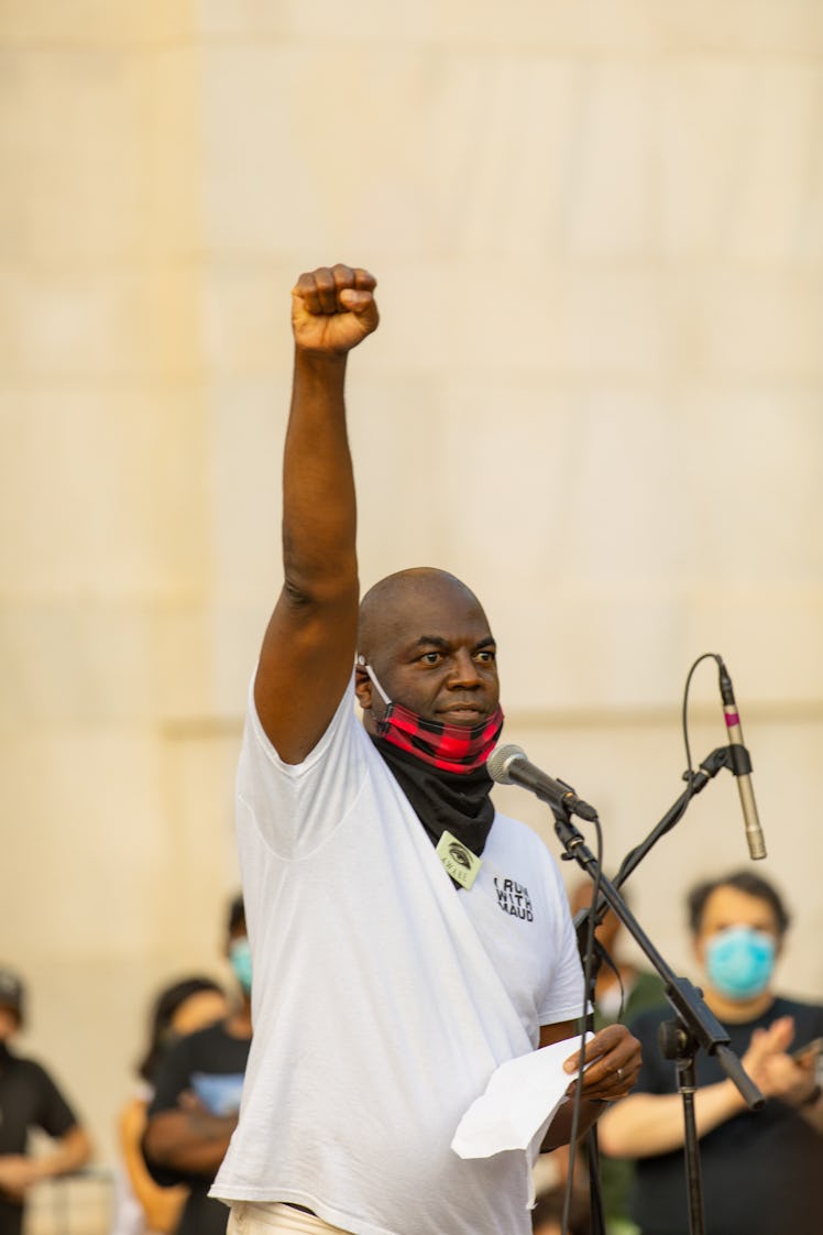 A man giving a speech in a white shirt and black-red face mask with one arm raised on the 4th of Jul...