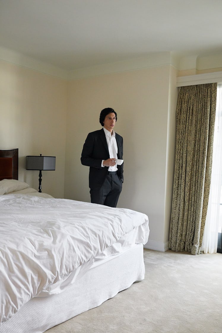 Adam Driver standing next to a bed while wearing a formal suit 