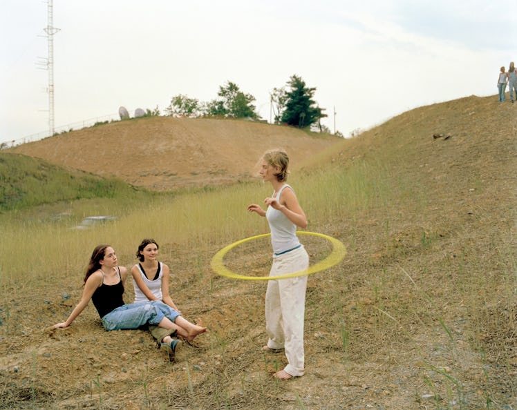 Girl wheeling yellow hoop around her waist while two other women are sitting on the grass besides he...