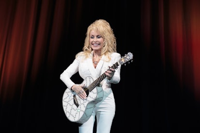 Dolly Parton in a white suit and a white guitar smiling and performing on a stage