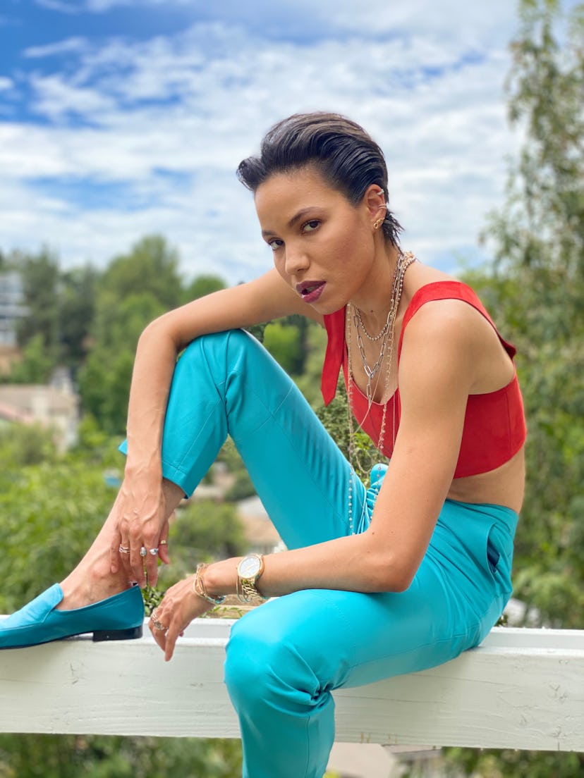 Jurnee Smollett in a red top, turquois pants and shoes sitting on a wooden beam 