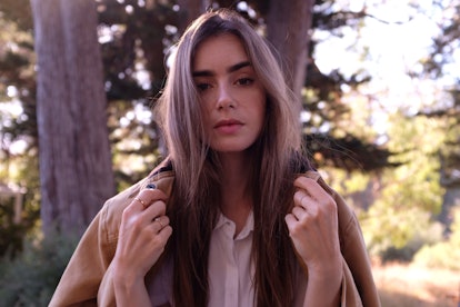 Lily Collins posing in a button up white shirt and brown jacket in the forest