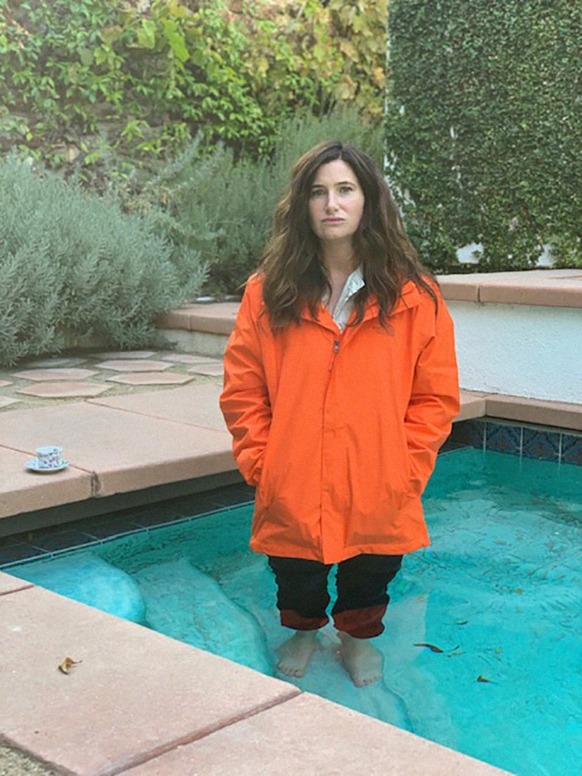 Kathryn Hahn standing in a pool, wearing an orange rain coat, black pants and a white button-up unde...