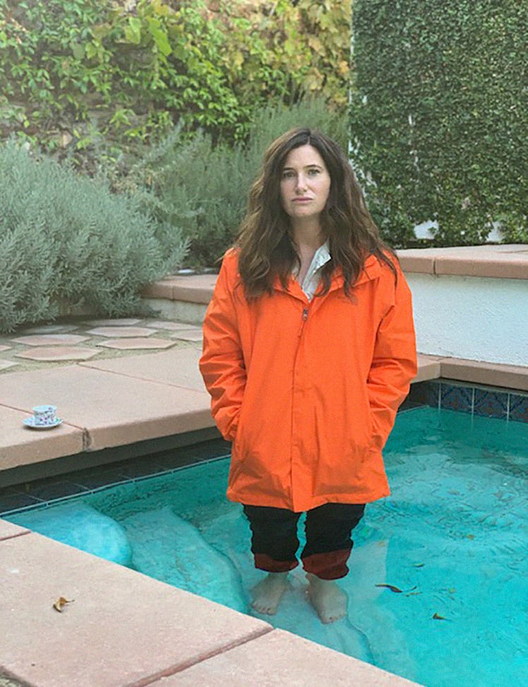 Kathryn Hahn standing in a pool, wearing an orange rain coat, black pants and a white button-up unde...