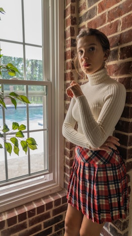 Stranger Thing’s Millie Bobby Brown posing in a white sweater and a red plaid skirt