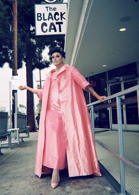 A drag performer in a long pink dress with a matching overcoat and shoes, leaning against a railing