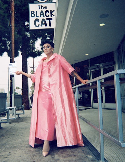A drag performer in a long pink dress with a matching overcoat and shoes, leaning against a railing