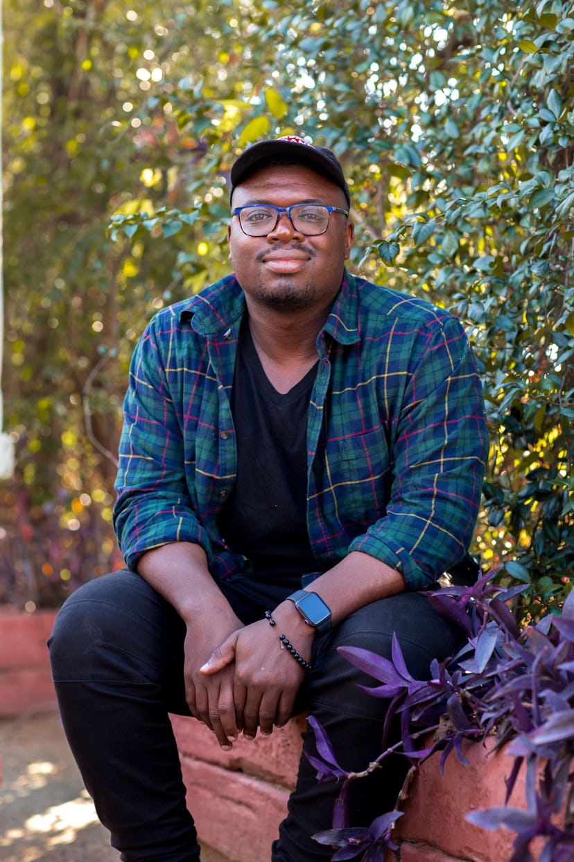 The writer Bryan Washington sitting in a garden while posing in a black shirt and pants, and a blue ...