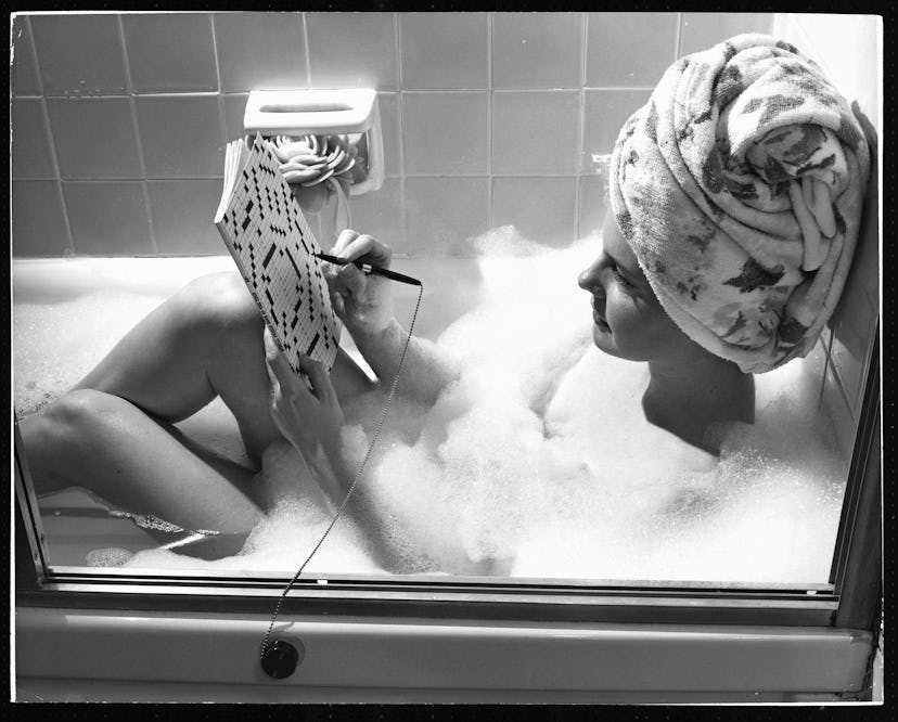 A woman doing her skincare routine in a bath while solving a crossword puzzle with her hair wrapped ...