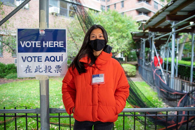 First-time poll worker Amanda Pong standing next to a "Vote Here" sign, in an orange puffer coat and...