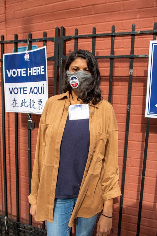 Vahni Kurra in a brown coat and jeans, standing next to a "Vote Here" sign
