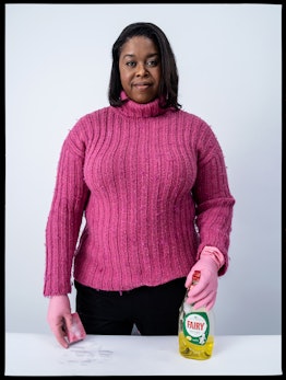Michelle Greenidge holding cleaning supplies in a pink knitted sweater, black pants, and pink cleani...
