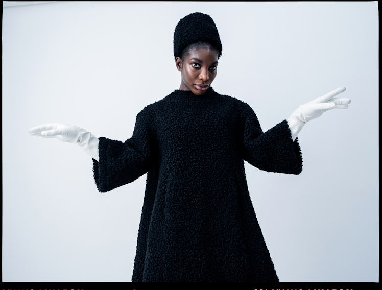 Michaela Coel posing in a plushy black dress and hat, with white gloves
