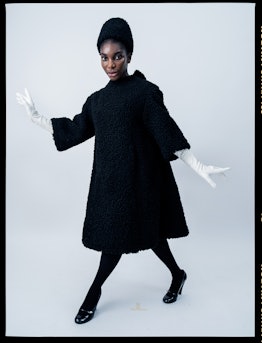 Michaela Coel wearing a plushy black dress and hat, black leggings, and heels, with white gloves