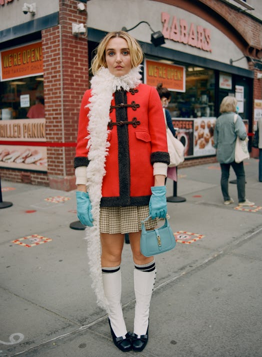 Chloe Fineman posing in a red Gucci coat with black details, a pleated skirt, and a white scarf pair...