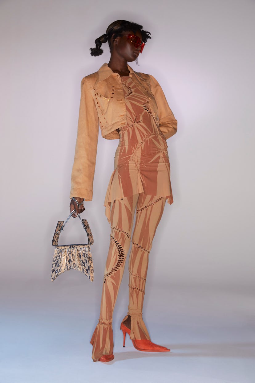 A model wearing a Charlotte Knowles peach and orange dress and tights, a peach jacket, and a snake-p...