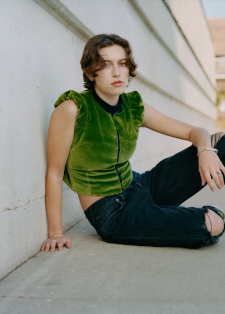 King Princess in an olive velvet vest and black trousers sitting and posing