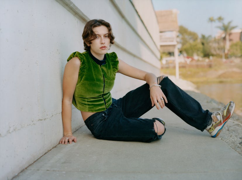 King Princess in an olive velvet vest and black trousers sitting and posing