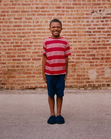 A boy in a red-white shirt and navy shorts smiling and posing for Matthew So’s series, Picture Day 2...