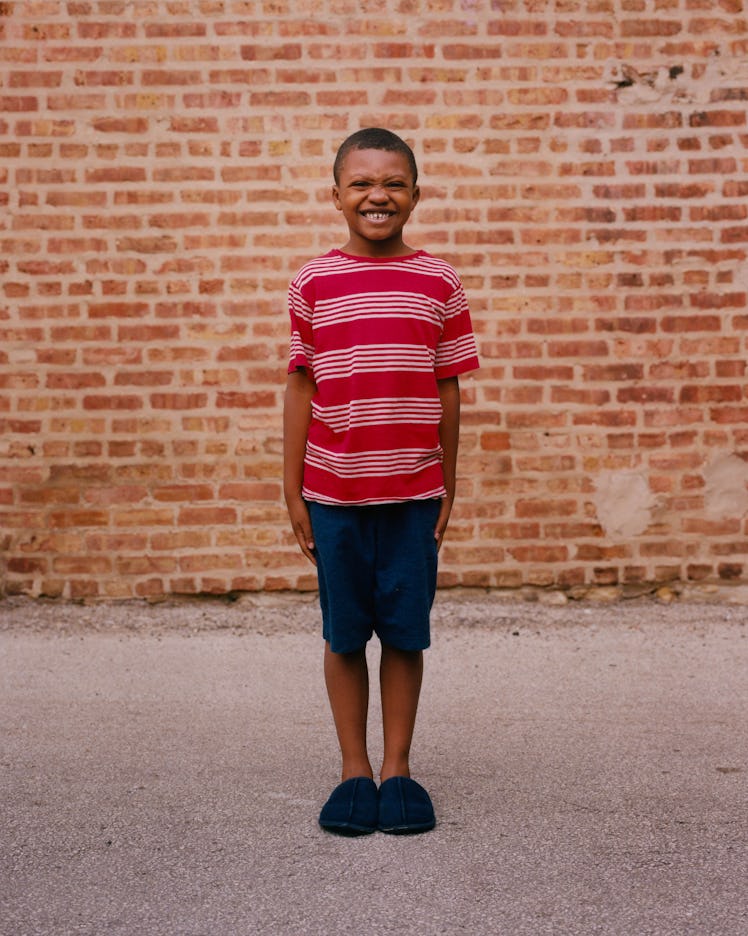 A boy in a red-white shirt and navy shorts smiling and posing for Matthew So’s series, Picture Day 2...