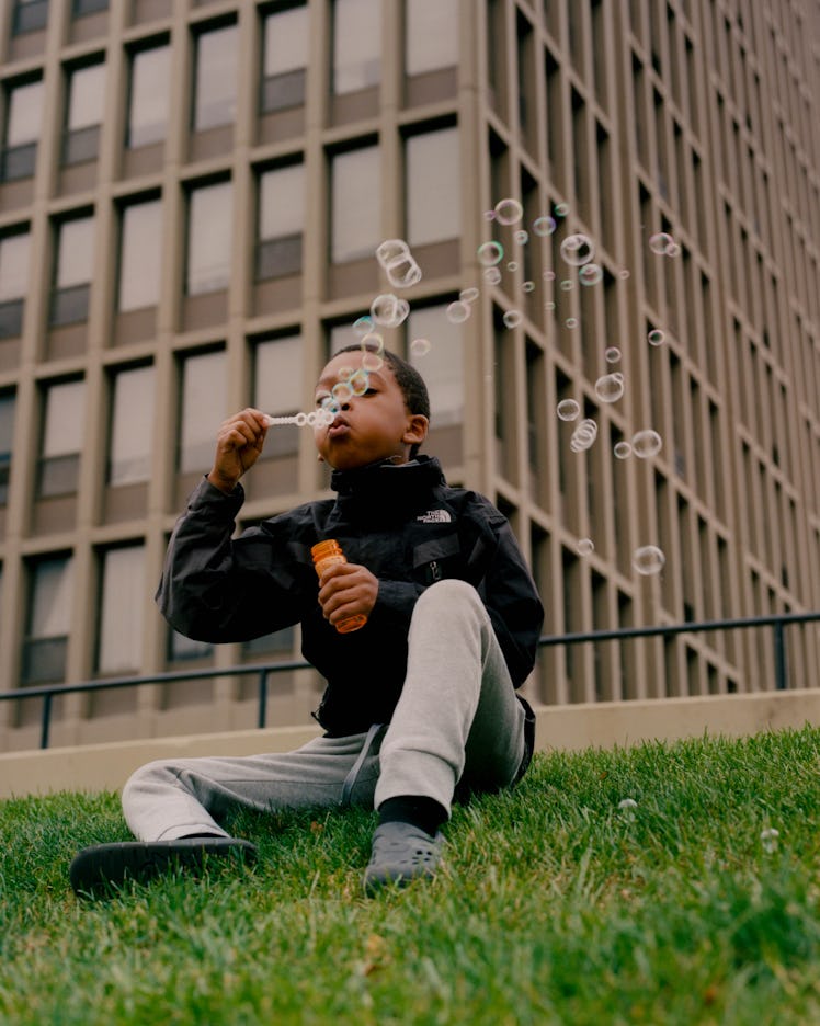 A boy in a black jacket and grey sweat pants blowing bubbles while posing for Matthew So’s series, P...