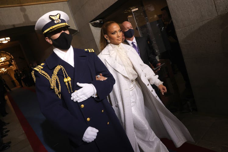 Jennifer Lopez in a white suit and a white coat escorted by a man in a black uniform