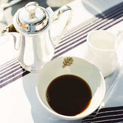 A coffee cup, half-filled with coffee and a metal coffee pot on a white table cloth with dark blue s...