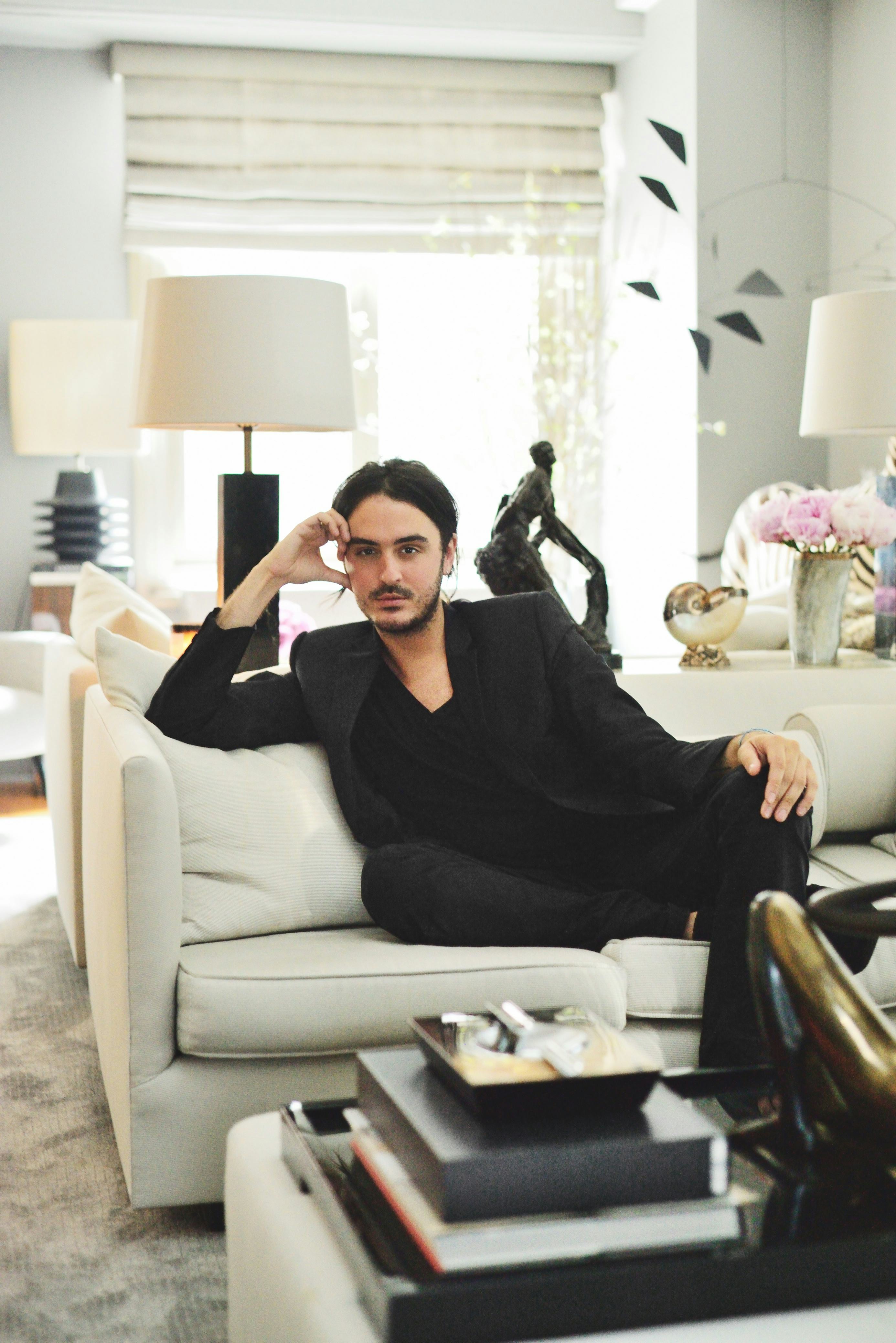 A man in a black suit sitting on a white couch in a glamourous home