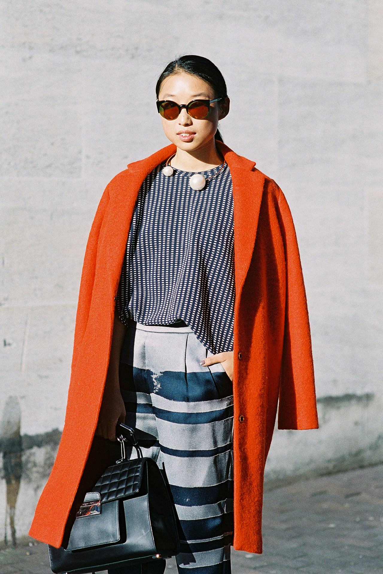 A woman in a stripy blouse with two large pearls, an orange coat and sunglasses