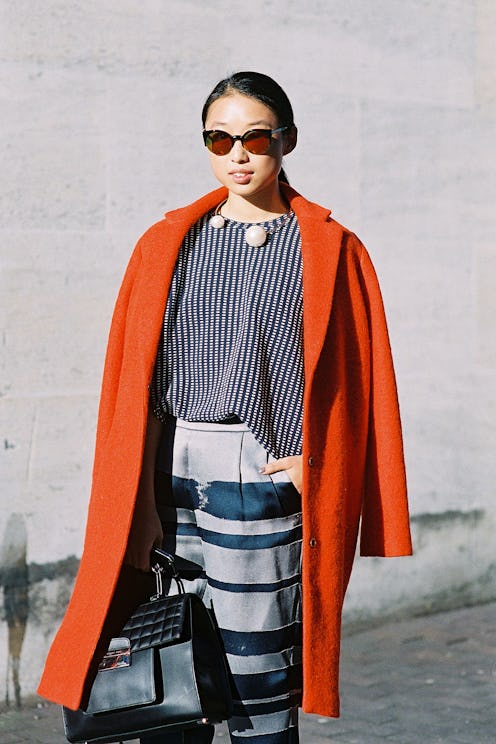 A woman in a stripy blouse with two large pearls, an orange coat and sunglasses