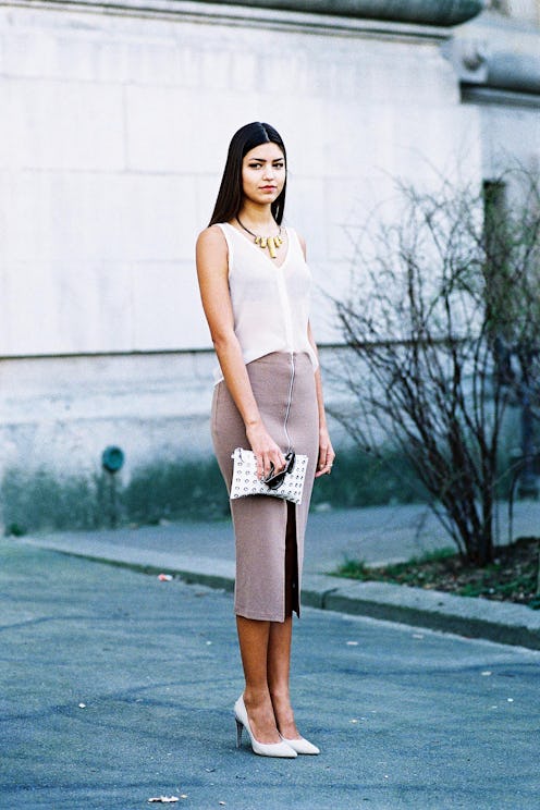 A brunette woman in a grey knit pencil skirt, white top, heels, and purse, standing