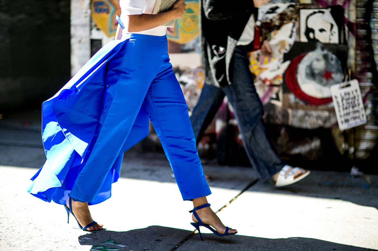 A woman walking in blue pants with material draping in the back and black sandal heels 