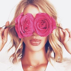 A woman holding two pink roses over her eyes while smiling and sticking out her tongue