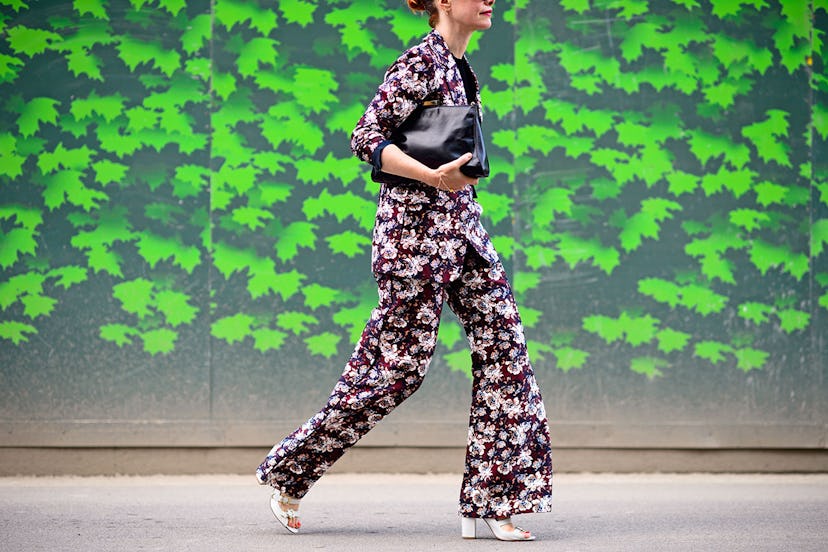 A woman walking in a floral suit, white sandal heels and a black bag 